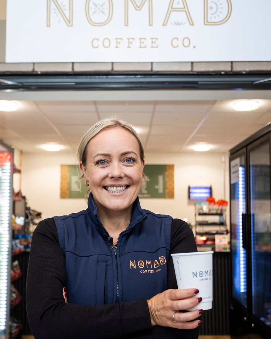 Cat Rainey in her navy Nomad gilet, arms folded with a Nomad cup, under the Parkway shop sign