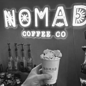 A hot chocolate piled with cream and marshmallows under the lit Nomad sign