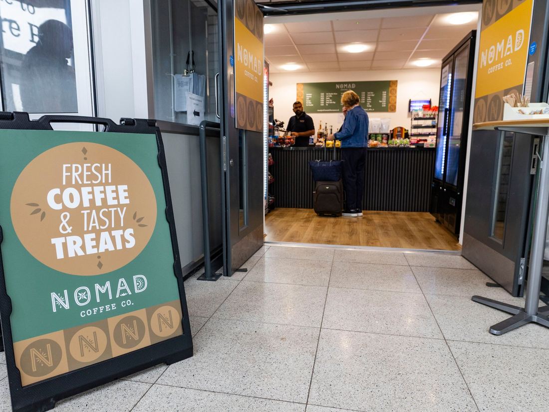 The green Fresh Coffee and Tasty Treats A-board outside the Nomad café at Worcestershire Parkway, with a traveller at the counter