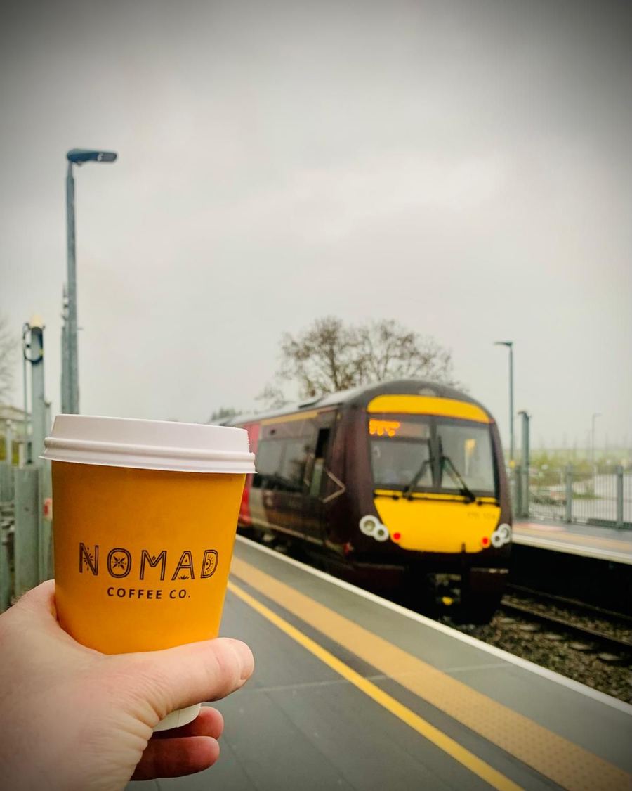 A mustard Nomad Coffee Co. cup held up on the platform as a train pulls in