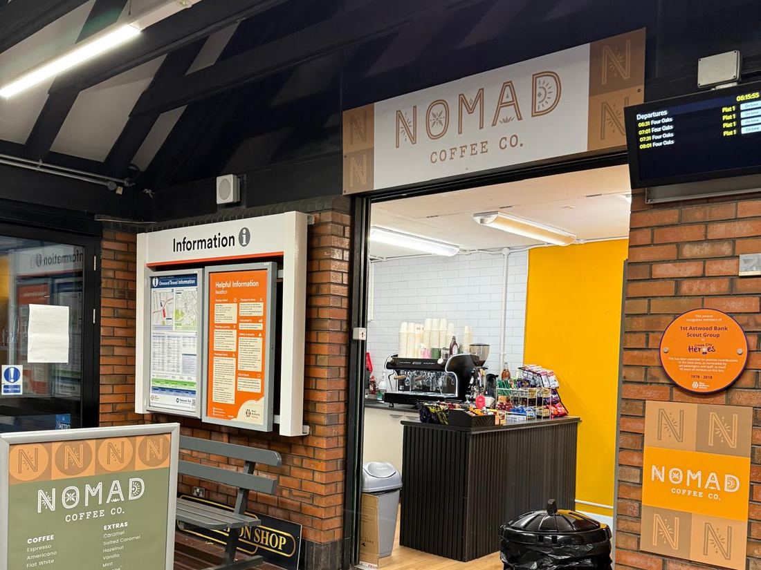 The Nomad shop at Redditch station beside the information board, with its green menu A-board outside and the departures screen showing early trains to Four Oaks